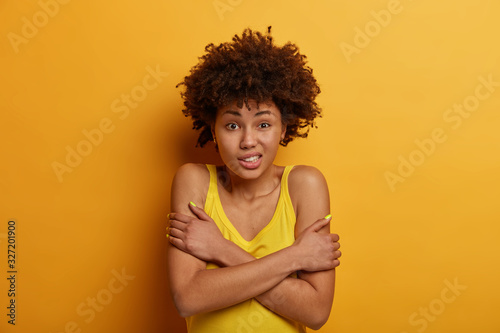Curly haired ethnic woman trembles from cold, crosses arms over body, clatters teeth, feels insecure and scared, wears casual yellow shirt, stands indoor, makes defensive pose, afraids of something