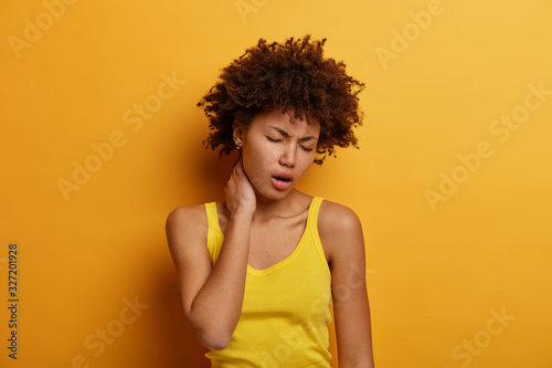 Upset fatigue curly young woman touches neck, suffers from painful feelings after sitting long time at computer, frowns face, wears yellow casual clothes, has problems with spine, poses indoor