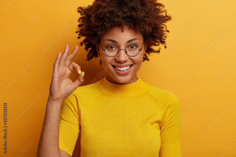 Pleased curly haired woman makes okay confirmation gesture, judges something, gives approval, says excellent, wears round spectacles and t shirt, isolated on yellow background, encourages not give up Pleased curly haired woman makes okay confirmation gesture, judges something, gives approval, says excellent, wears round spectacles and t shirt, isolated on yellow background, encourages not give up