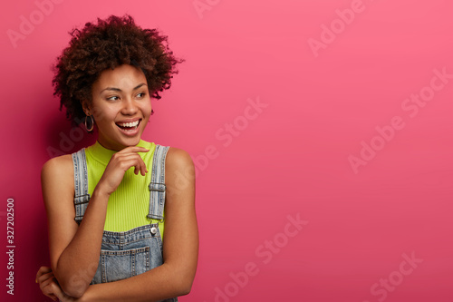 Half length shot of dark skinned girl keeps gaze aside, holds hand under chin, wears denim overalls, being in high spirit, poses over rosy studio background, copy space aside for your promotion