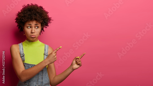 Disappointed upset woman with Afro hair indicates aside with gloomy expression, shows something unpleasant, dressed casually, stands against bright pink background, displeased with high prices © Wayhome Studio