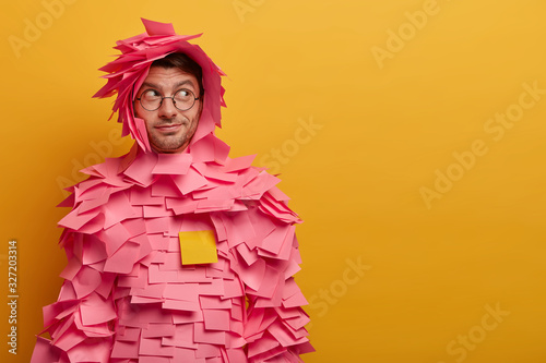 Indoor shot of male advertiser or manager covered with sticky adhesive notes, looks aside as notices something interesting, poses against yellow background, free space for your advertising content