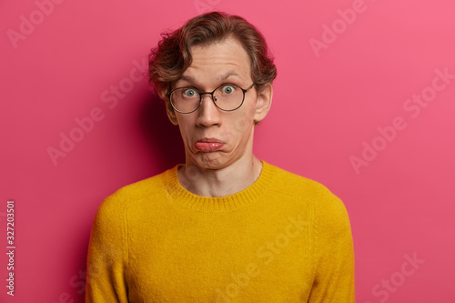 Stunned emotional guy purses lips and looks with wonder at camera  feel puzzled and surprised about hearing some information  wears yellow sweater and transparent glasses  isolated on pink wall