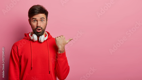Tableau sur toile Studio shot of amazed bearded young man points thumb aside, has shocked expressi