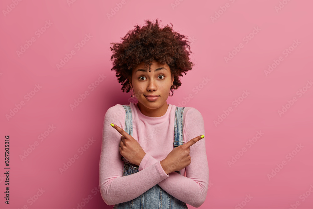 Hesitant clueless woman with Afro hair thinks what to order, crosses arms over body and points sideways, wears turtleneck, has surprised gaze at camera, tries to make choice, shows two variants Hesitant clueless woman with Afro hair thinks what to order, crosses arms over body and points sideways, wears turtleneck, has surprised gaze at camera, tries to make choice, shows two variants