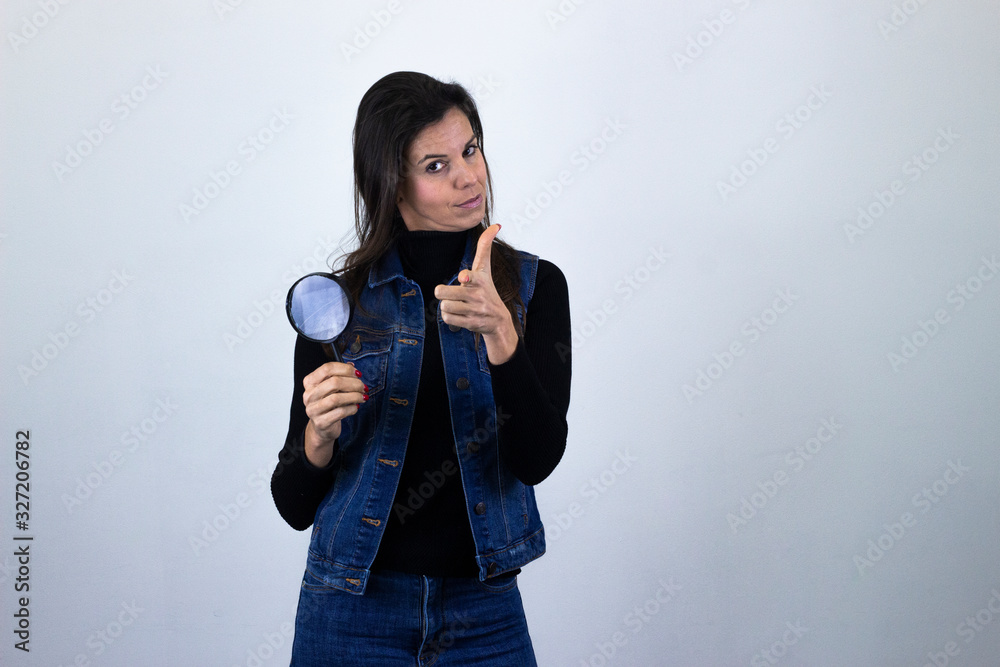 Fototapeta premium Portrait of confident ambitious successful attractive caucasian middle age woman model with magnifying glass looking directly at camera, showing direction with one forefinger, isolated on gray backgro
