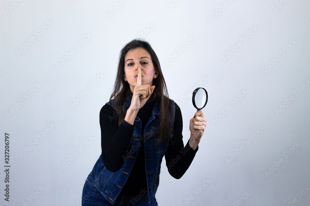 Portrait of attractive caucasian middle age woman with magnifying glass holding index finger near his lips, silence, isolated on gray background studio shot, black sweater, denim jacket, jeans, dark a Portrait of attractive caucasian middle age woman with magnifying glass holding index finger near his lips, silence, isolated on gray background studio shot, black sweater, denim jacket, jeans, dark a