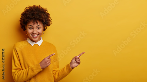 Cheerful pretty woman with Afro hair points at copy space, shows recommendation, suggests going this direction or promo, wears neat sweater, isolated over yellow background, makes good choice