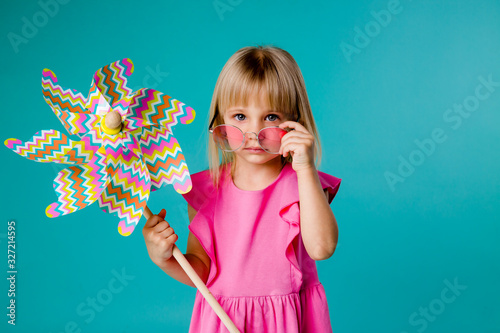 child girl blonde smiling in sunglasses in a pink dress holding a windmill on a blue background of isolate. the concept of the summer, space for text