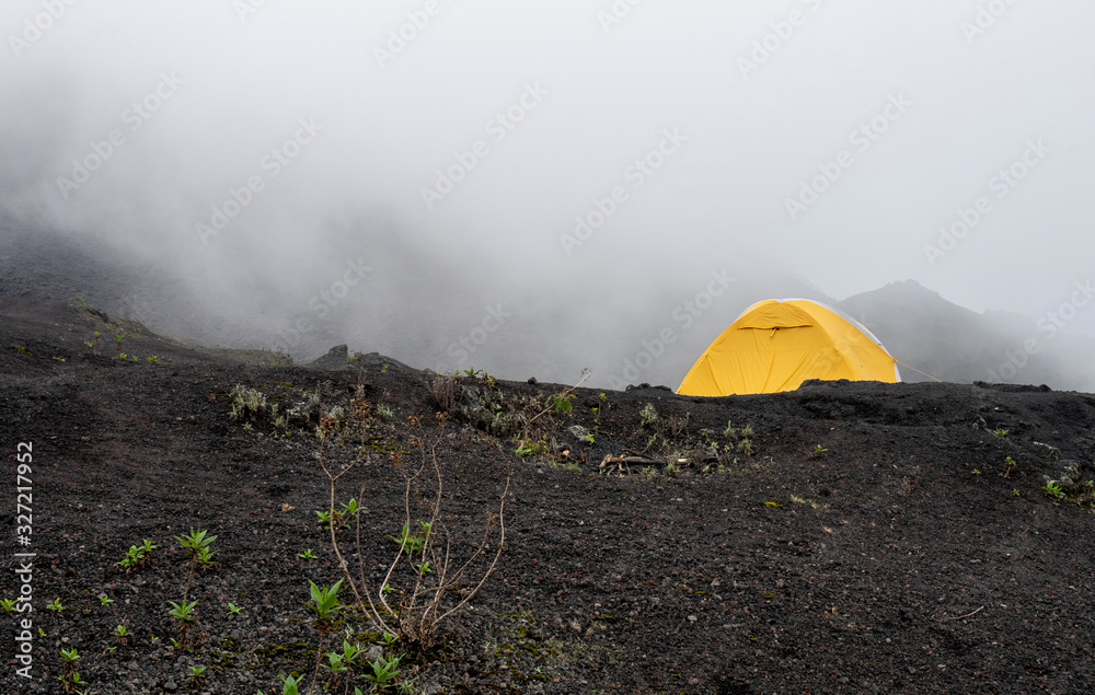 Yellow camping tent covered in fog at the top of Pacaya volcano in Guatemala, black rock floor and some vegetation