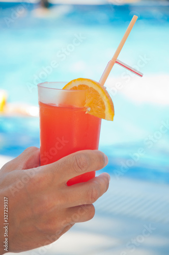 Fresh cocktail with orange slice and plastic tube in man's hand on blue pool water background