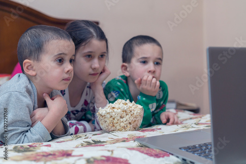 Children looking at laptop with cartoons showing before sleeping in bed. Three kids in bed watching a movie on a laptop and eating popcorn. toned