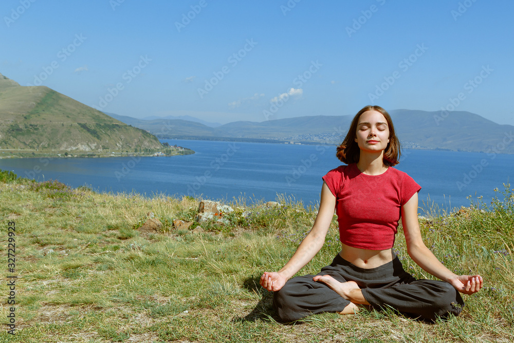 Young woman doing yoga and meditating in mountain landscape. Concentrated and contented female sitting with closed eyes in lotus position at grass with lake behind in sunny day 