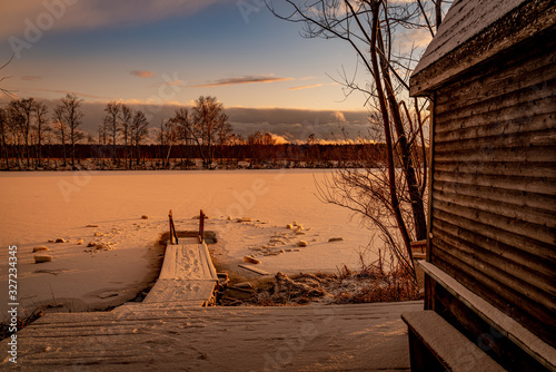 Beautiful view of old wooden bathhouse on the shore of a frozen river with the pier at sunset. Travel destination Russia