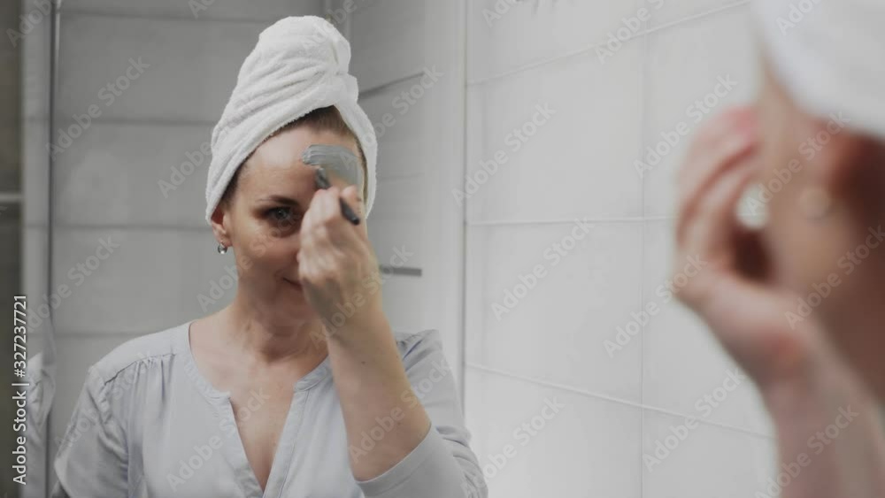Adult woman with a towel on her head applying clay mask on her face looking at the camera Adult woman with a towel on her head applying clay mask on her face looking at the camera