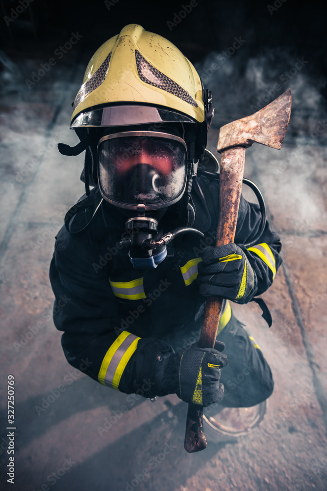 Obraz premium Portrait of a female firefighter while holding an axe and wearing an oxygen mask indoors surrounded by smoke.
