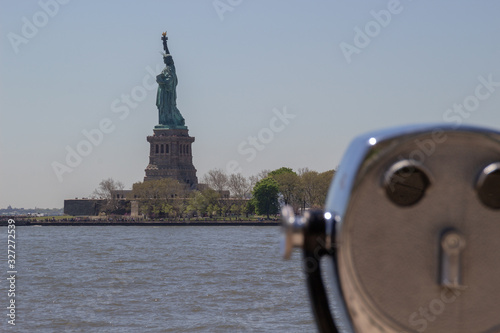 Tourist binoculars on Ellis Island with Statue of Liberty visible in the blurred background. There is a blue sky and water visible. The metal shows age and is burnished © Dmitriy