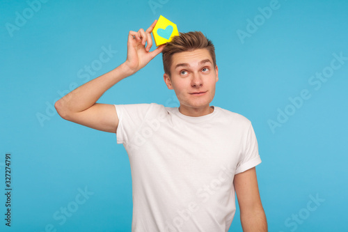 Dreaming of home purchase. Positive thoughtful man in t-shirt holding small paper house on head, planning to buy apartment, looking up with pensive expression. studio shot isolated on blue background