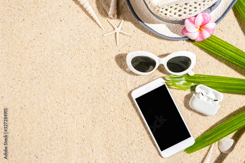Summer holiday concept with white hat, sunglasses, phone, headphone, sea shells and starfish on sand background with green palm branch, copy space, flat lay