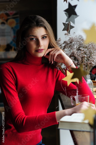 Beautiful girl in a red sweater at the window in a cafe with a cup of tea and a book