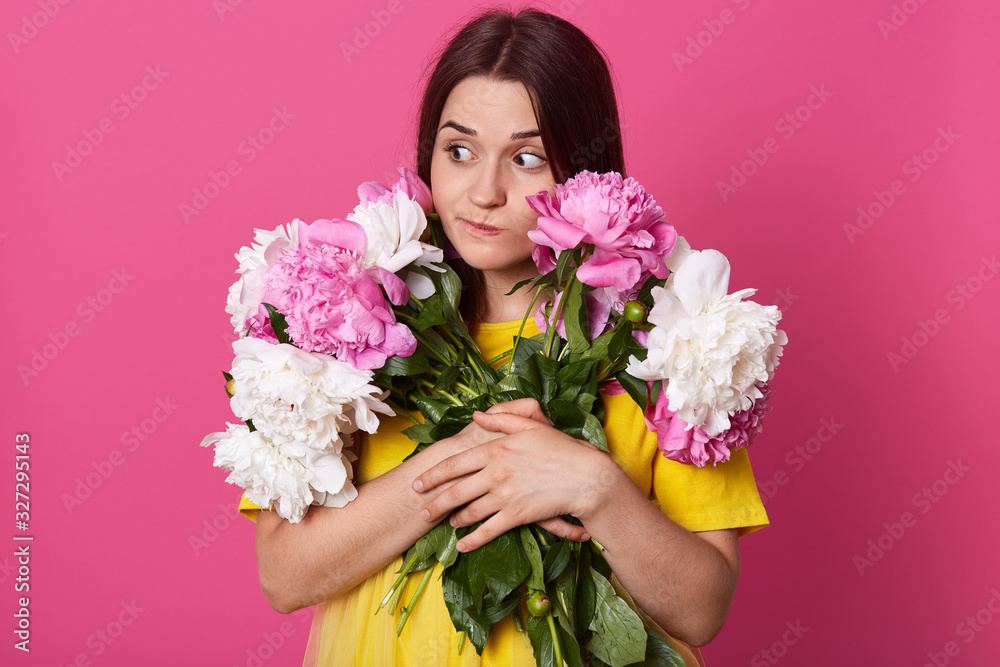 Close up portrait of pencive brunette woman embracing perfect bouquet of peony flowers, posing isolated over rosy background, female looking aside with thoufhtful and cunning facial expression.