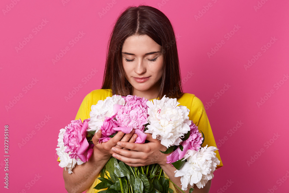 Indoor shot of beautiful dark haired female embracing big bouquet with pink and white peony flowers, has gentle look, posing isolated over pink studio background. Women's Day, 8 march concept. Indoor shot of beautiful dark haired female embracing big bouquet with pink and white peony flowers, has gentle look, posing isolated over pink studio background. Women's Day, 8 march concept.