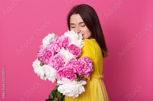 Portrait of beautiful brunette girl standing with closed eyes, looking gentle, brunette woman wearing yellow dress, model embracing pink and white peony flowers, enjoying her presant from boyfriend. photo