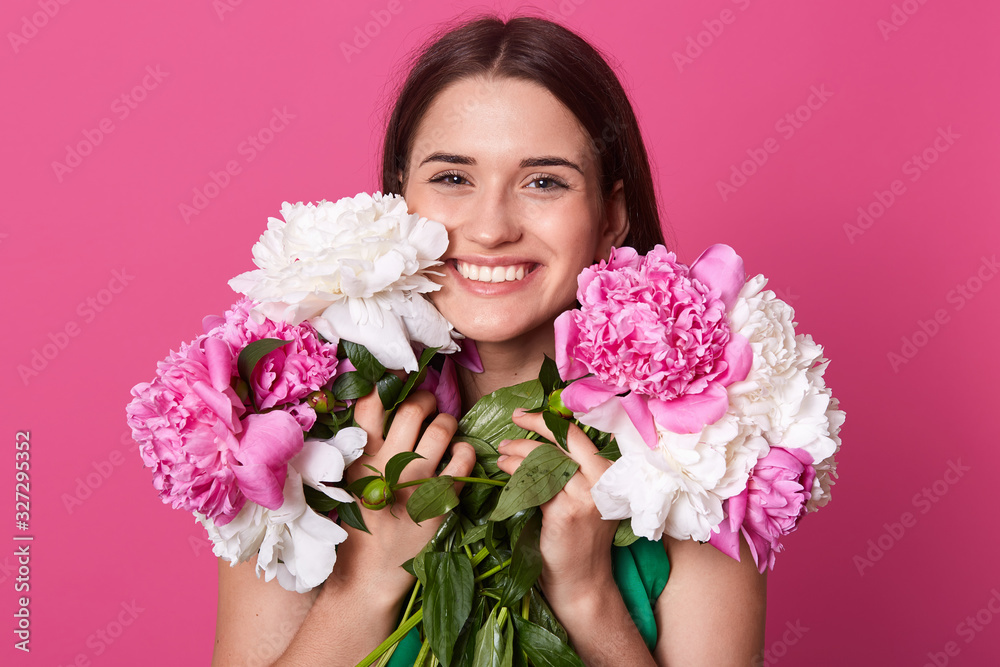 Studio shot of charismatic female standing isolated over rosy background, looking directly at camera and laughing happily ,holding big bouquet of white and pink peonies, female expressing happyness.