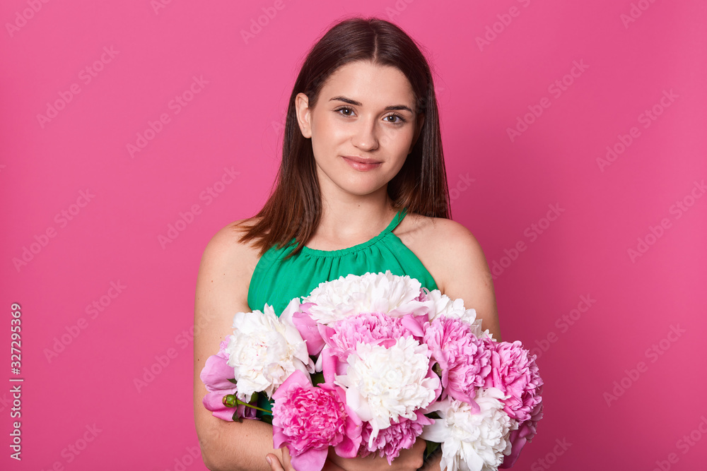 Beautiful woman wearing green sundress, holding bouquet of gentle peonies. Adorable lady stands miling and looking at camera, looks happy, embracing flowers fron her boyfriend for 8 march. Women's day