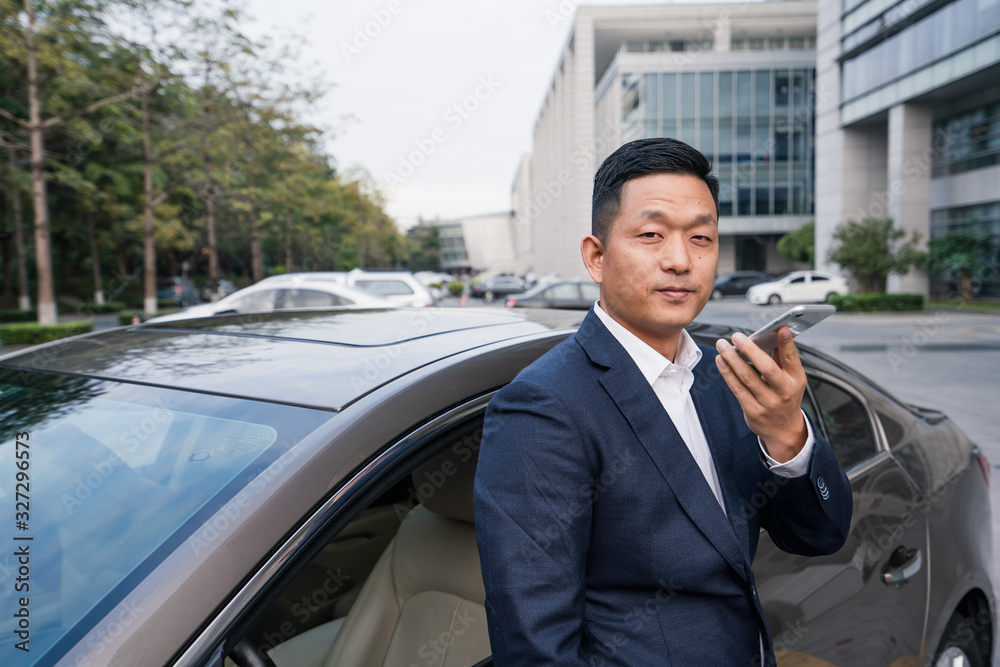 Asian man in front of the office building is on the phone beside the car Asian man in front of the office building is on the phone beside the car