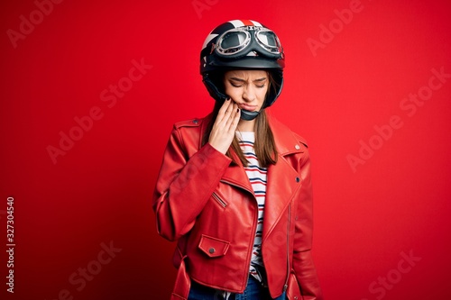 Young beautiful brunette motocyclist woman wearing motorcycle helmet and red jacket touching mouth with hand with painful expression because of toothache or dental illness on teeth. Dentist © Krakenimages.com