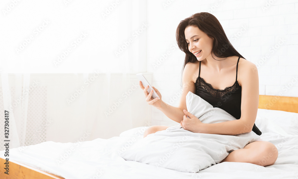 Happy woman sitting in bed, smiling and reading text message Happy woman sitting in bed, smiling and reading text message