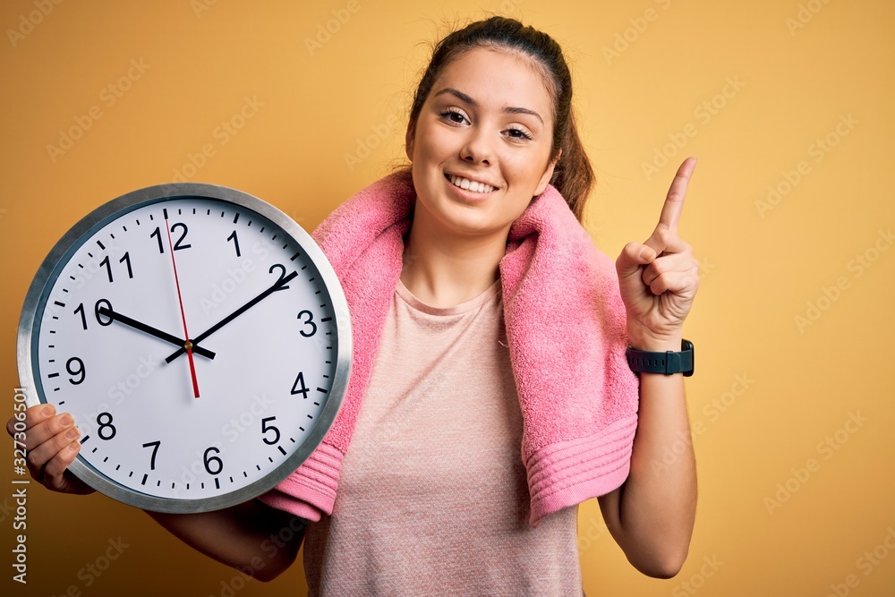 Young beautiful brunette sportswoman wearing towel doing countdown using big clock surprised with an idea or question pointing finger with happy face, number one Young beautiful brunette sportswoman wearing towel doing countdown using big clock surprised with an idea or question pointing finger with happy face, number one