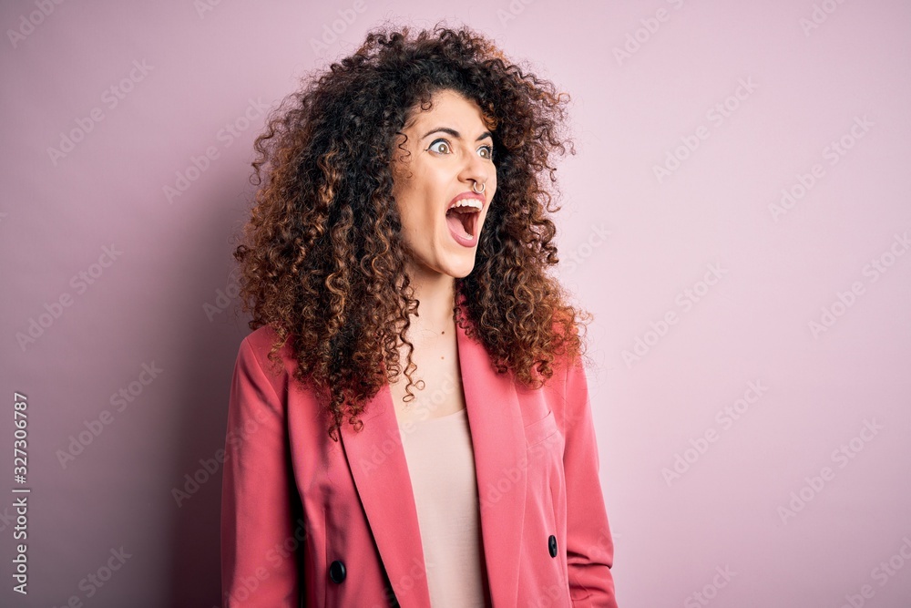 Young beautiful businesswoman with curly hair and piercing wearing elegant jacket angry and mad screaming frustrated and furious, shouting with anger. Rage and aggressive concept. Young beautiful businesswoman with curly hair and piercing wearing elegant jacket angry and mad screaming frustrated and furious, shouting with anger. Rage and aggressive concept.