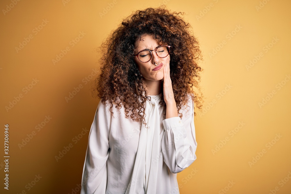 Young beautiful brunette woman with curly hair and piercing wearing shirt and glasses touching mouth with hand with painful expression because of toothache or dental illness on teeth. Dentist Young beautiful brunette woman with curly hair and piercing wearing shirt and glasses touching mouth with hand with painful expression because of toothache or dental illness on teeth. Dentist