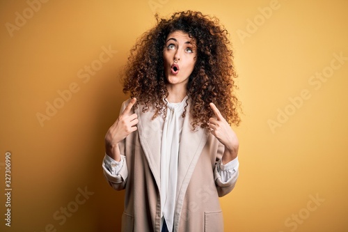 Young beautiful brunette woman with curly hair and piercing wearing casual t-shirt and diadem amazed and surprised looking up and pointing with fingers and raised arms.
