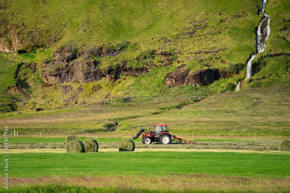 Obraz premium Red Tractor collecting grass on green field with beautiful river going by the mountain on background