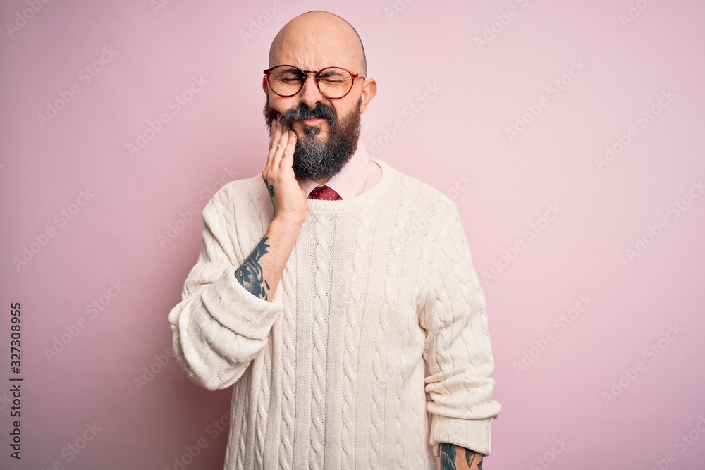 Handsome bald man with beard and tattoo wearing glasses and sweater over pink background touching mouth with hand with painful expression because of toothache or dental illness on teeth. Dentist