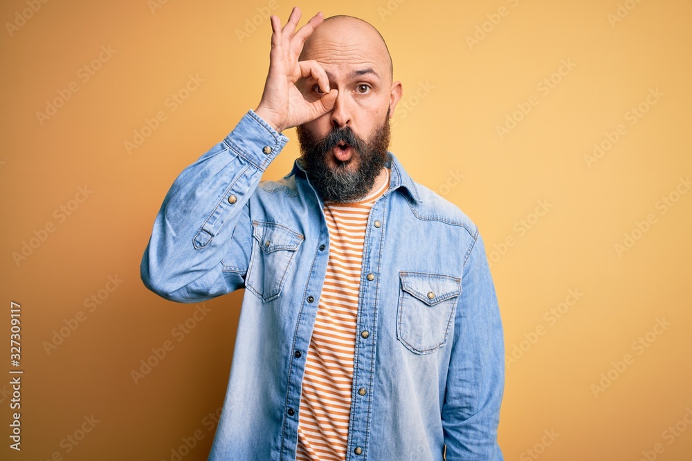 Obraz premium Handsome bald man with beard wearing casual denim jacket and striped t-shirt doing ok gesture shocked with surprised face, eye looking through fingers. Unbelieving expression.