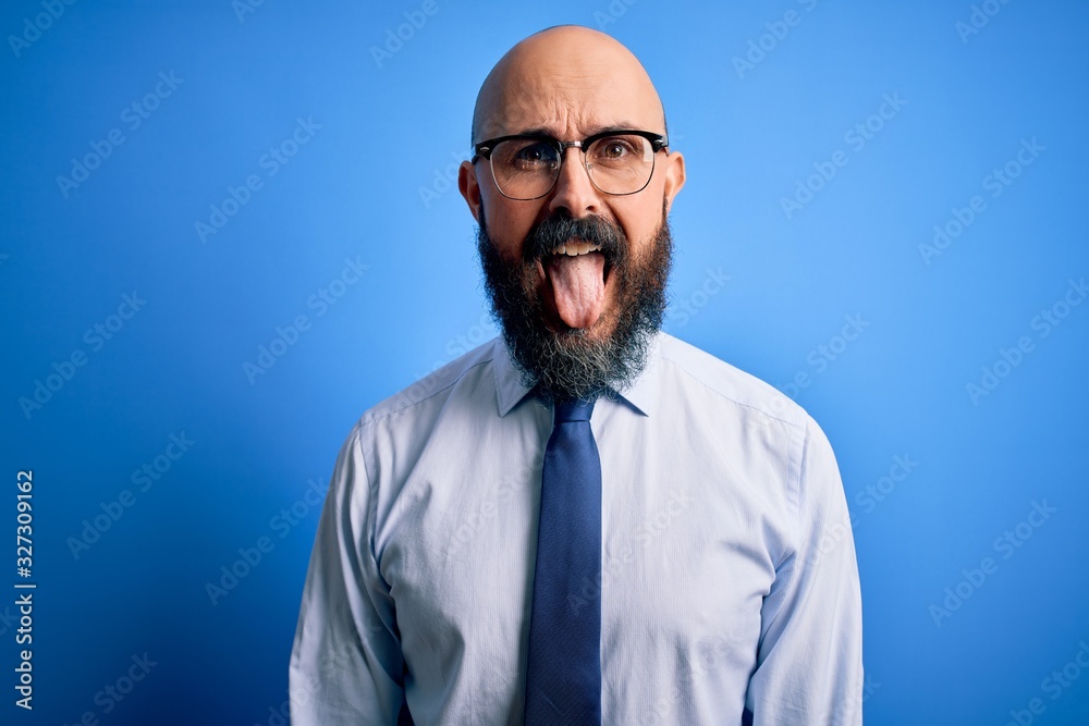 Handsome business bald man with beard wearing elegant tie and glasses over blue background sticking tongue out happy with funny expression. Emotion concept.