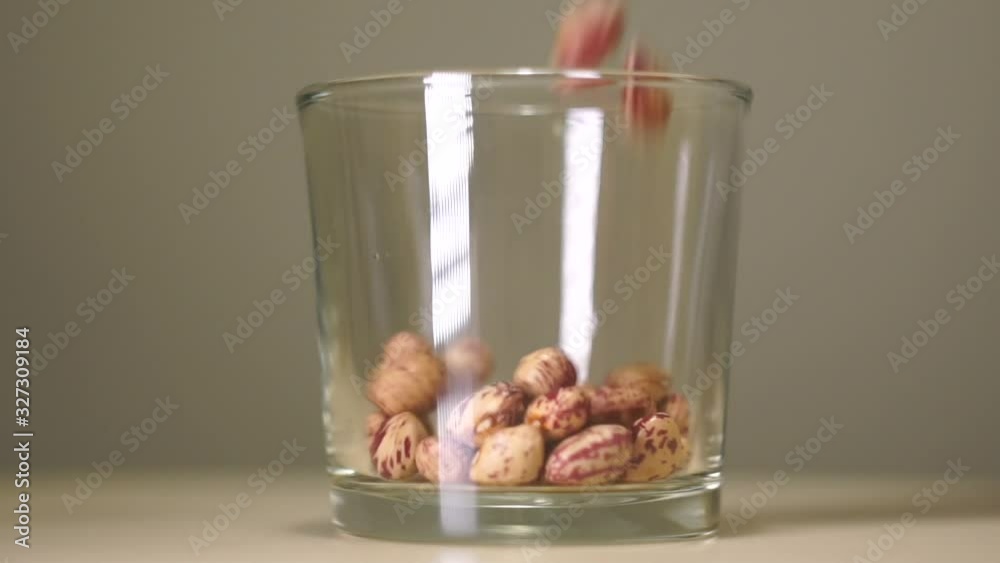 Raw pinto beans (the most popular beans in the United States and northwestern Mexico) in transparent glass with white reflection. Cereals pouring into an empty glass on a pastel background, close-up.