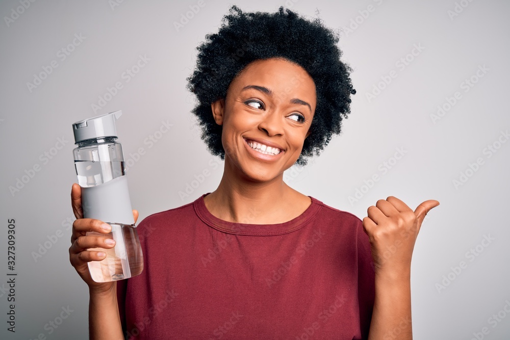 Young African American afro woman with curly hair drinking bottle of water for refreshment pointing and showing with thumb up to the side with happy face smiling Young African American afro woman with curly hair drinking bottle of water for refreshment pointing and showing with thumb up to the side with happy face smiling
