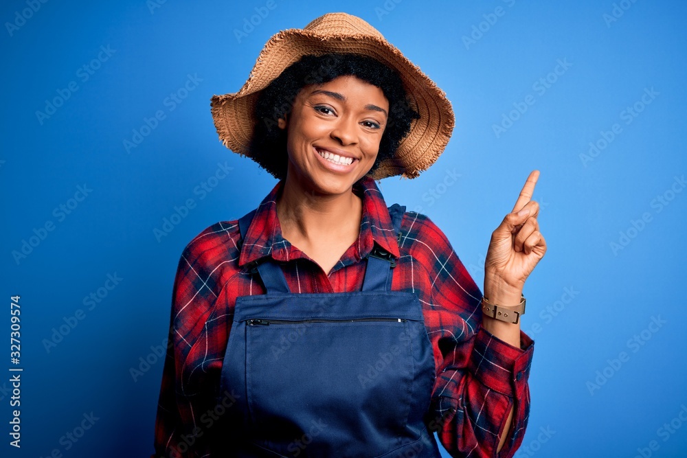 Young African American afro farmer woman with curly hair wearing apron and hat with a big smile on face, pointing with hand and finger to the side looking at the camera. Young African American afro farmer woman with curly hair wearing apron and hat with a big smile on face, pointing with hand and finger to the side looking at the camera.