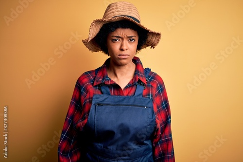 Young African American afro farmer woman with curly hair wearing apron and hat skeptic and nervous, frowning upset because of problem. Negative person.
