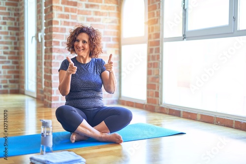 Middle age beautiful sportswoman wearing sportswear sitting on mat practicing yoga at home pointing fingers to camera with happy and funny face. Good energy and vibes.