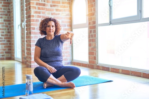 Middle age beautiful sportswoman wearing sportswear sitting on mat practicing yoga at home looking unhappy and angry showing rejection and negative with thumbs down gesture. Bad expression.