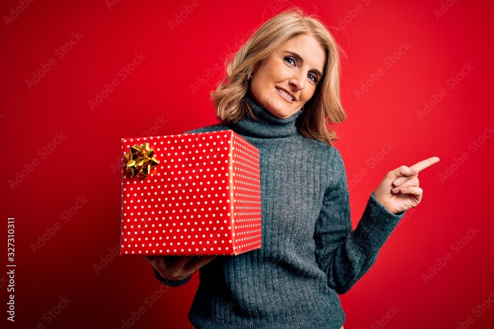 Middle age beautiful blonde woman holding birthday gift over isolated red background very happy pointing with hand and finger to the side Middle age beautiful blonde woman holding birthday gift over isolated red background very happy pointing with hand and finger to the side