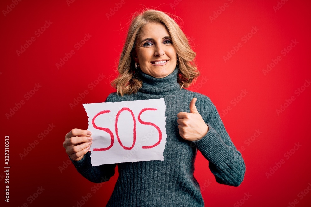 Middle age beautiful blonde woman holding paper with sos message over red bakground happy with big smile doing ok sign, thumb up with fingers, excellent sign Middle age beautiful blonde woman holding paper with sos message over red bakground happy with big smile doing ok sign, thumb up with fingers, excellent sign