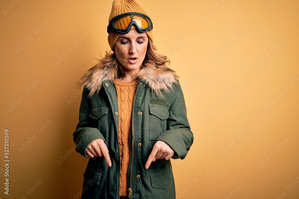 Middle age beautiful blonde skier woman wearing snow sportwear and ski goggles Pointing down with fingers showing advertisement, surprised face and open mouth