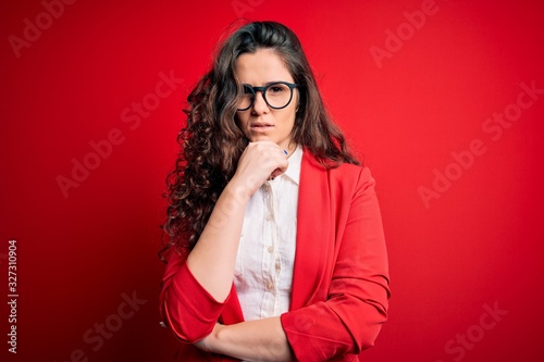 Young beautiful woman with curly hair wearing jacket and glasses over red background with hand on chin thinking about question, pensive expression. Smiling with thoughtful face. Doubt concept.